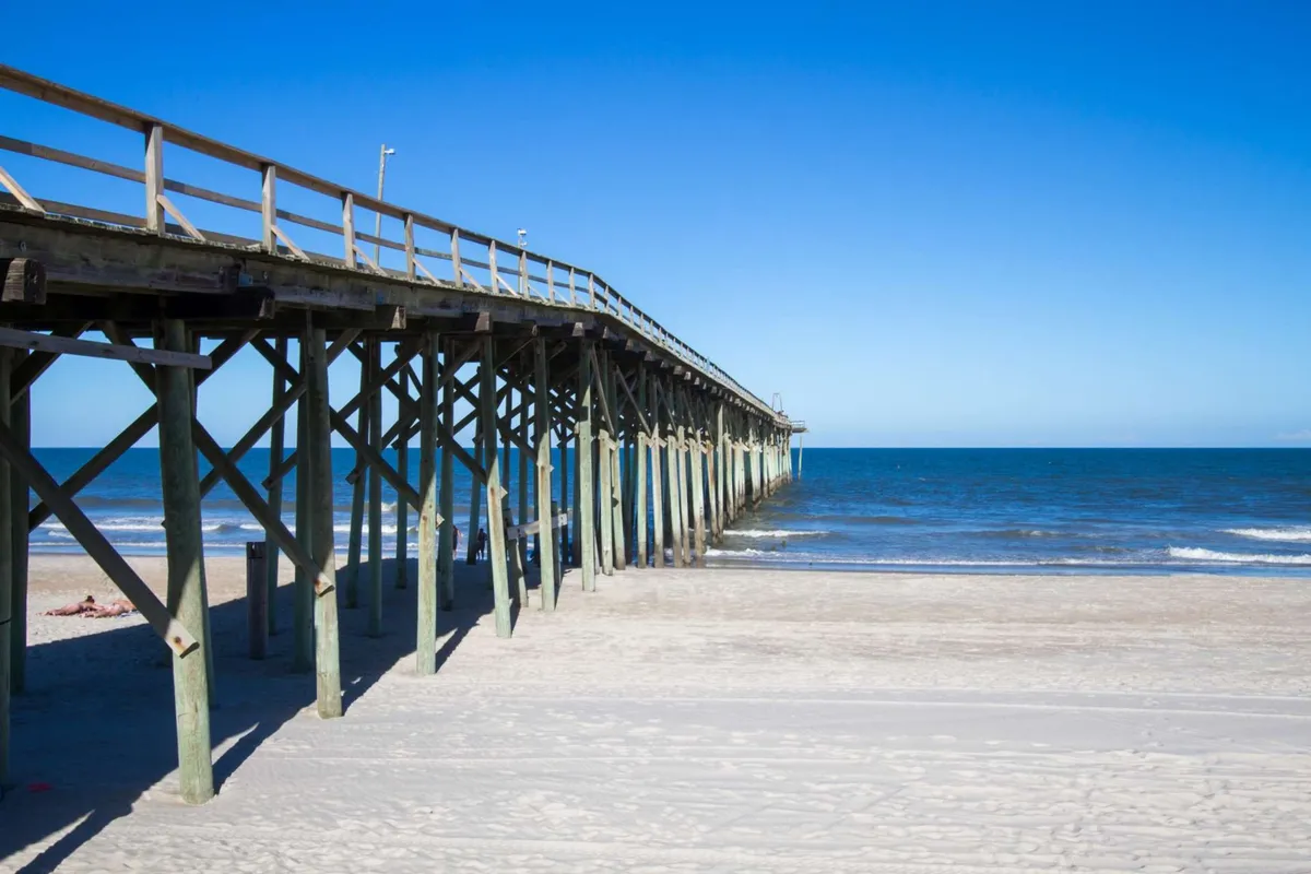 A classic wooden pier extends over the ocean with gentle waves lapping against the shore. The sandy beach in the foreground is open for activities, while the pier itself is a hub for fishing, strolls, and enjoying expansive water views.