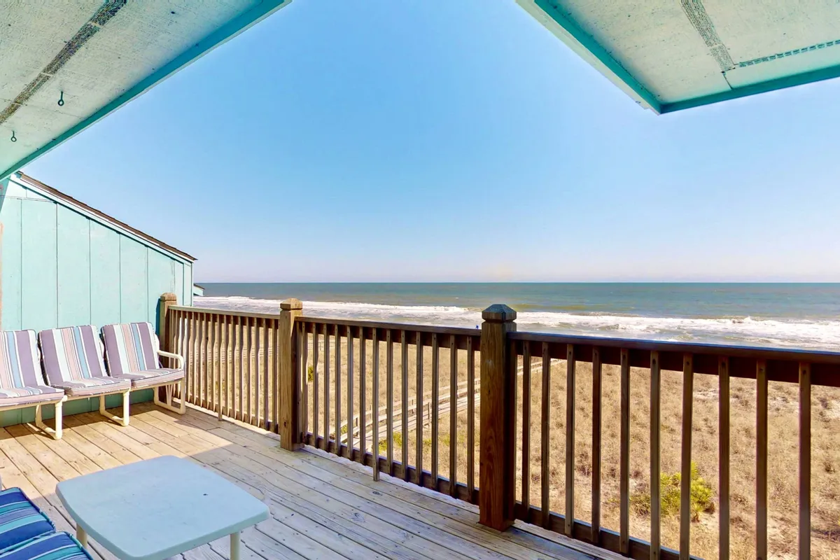 A tranquil exterior view featuring a charming blue cottage with deck seating that overlooks the ocean. The deck is an inviting spot for sunbathing, reading, or enjoying coffee at sunrise, with the serene ocean stretching out beyond.