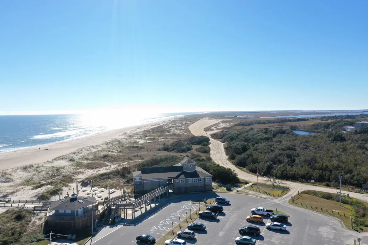 This image shows a sweeping view of the ocean, a sandy beach area, a wooden boardwalk leading towards the water, a parking lot, and natural coastal landscape. The inviting open spaces suggest easy access to the beach and opportunities for walks, fishing, and relaxing in a natural setting.