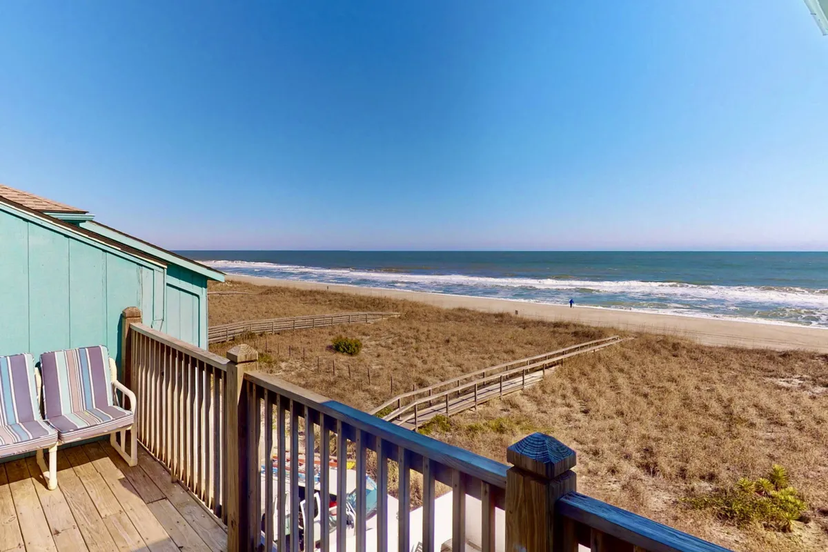 The image showcases a peaceful beachfront view from a private wooden deck equipped with inviting chairs. The deck overlooks the pristine sands and rolling waves of Carolina Beach, with the ocean stretching out to the horizon. The early morning sunlight or late afternoon glow suggests the perfect spot for relaxing, watching the sunrise, or enjoying an evening drink as the sun sets. The atmosphere is tranquil and promises a rejuvenating escape.