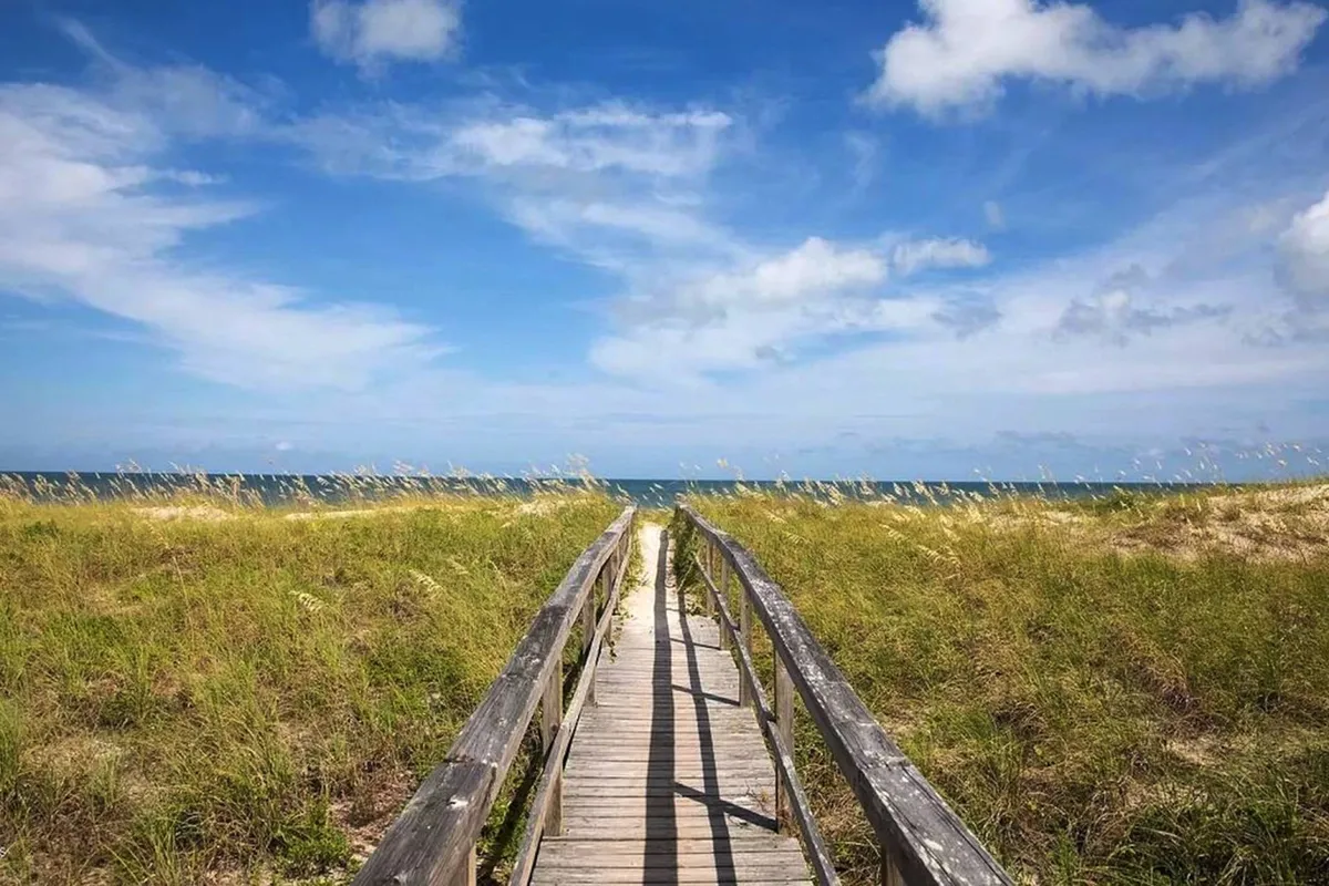 A serene wooden boardwalk stretching through tall grass toward the open ocean under a vivid blue sky. The scene invites you to imagine walking directly from the community to the beach, capturing the tranquility and direct beach access the property offers.