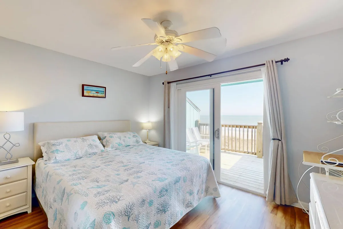 A serene bedroom with a king bed dressed in soft linens, a lamp and decorative painting adding warmth, and a sliding glass door leading to a private balcony with ocean views. The atmosphere is calm, suggesting a perfect space for relaxation after a day at the beach.