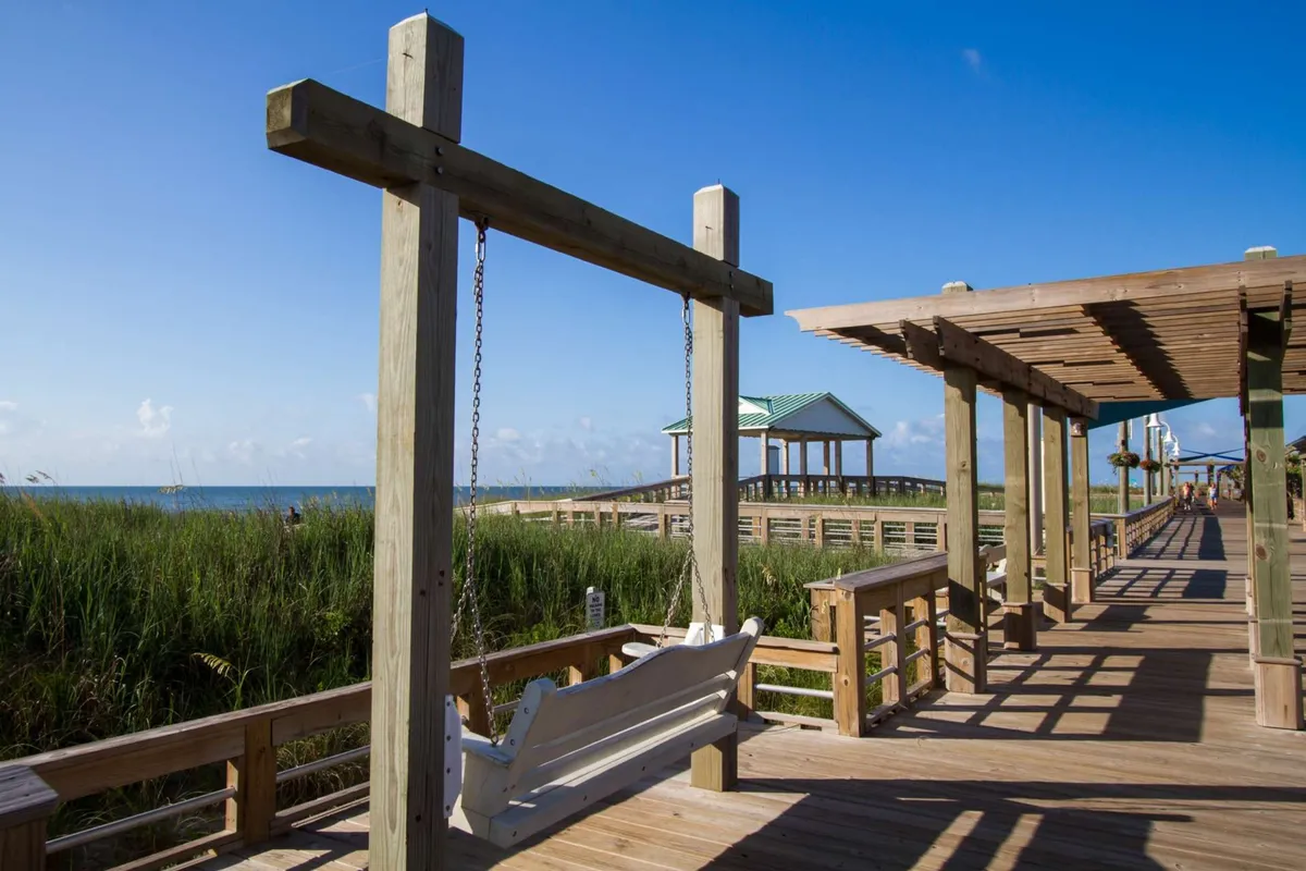 A scenic view of a wooden boardwalk with a swing overlooking the ocean and a pavilion in the background. The image evokes relaxation, with the swing offering a perfect spot to enjoy the ocean breeze and panoramic views.