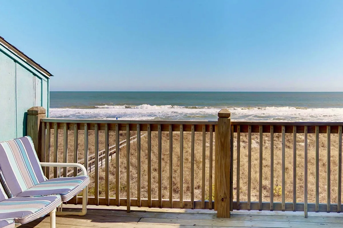 An elevated exterior view from the deck, featuring striped chairs, a sturdy railing, and an expansive sightline to the ocean and sandy beach beyond. The tranquil scene is set for relaxation, socializing, and enjoying the sights and sounds of the sea.