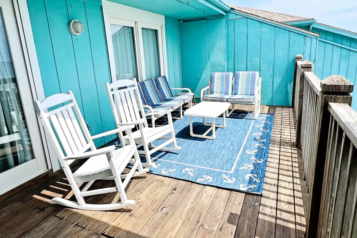 An inviting balcony set with white rocking chairs and striped lounge chairs atop a blue area rug, positioned to take in the ocean scenery. The area feels private yet open, perfect for morning coffee or evening cocktails with friends and family.