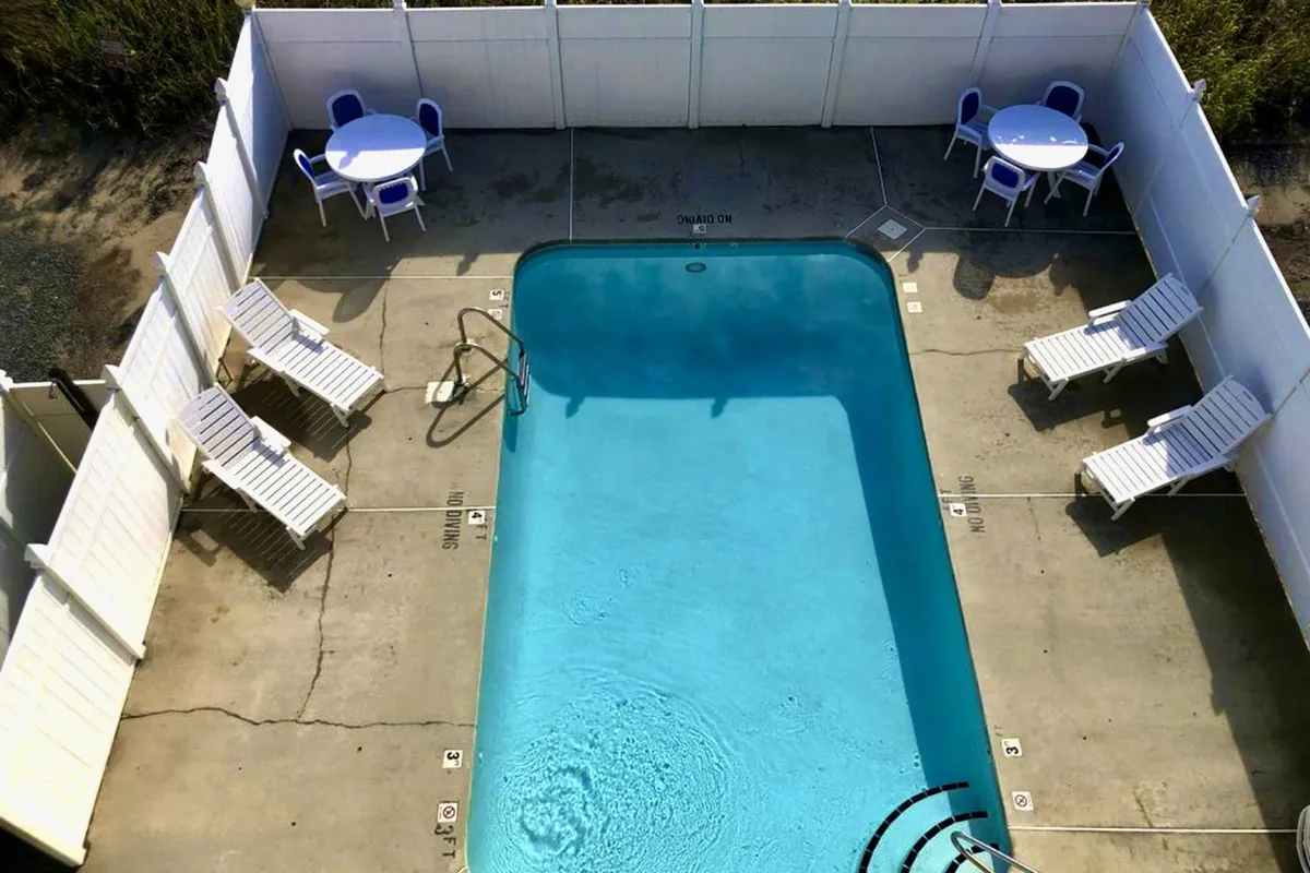 A bright, inviting community swimming pool surrounded by lounge chairs and an outdoor table with chairs, ideal for relaxing or socializing. The pool area is framed by lush greenery and captures the resort-style amenity that is a highlight of the community.