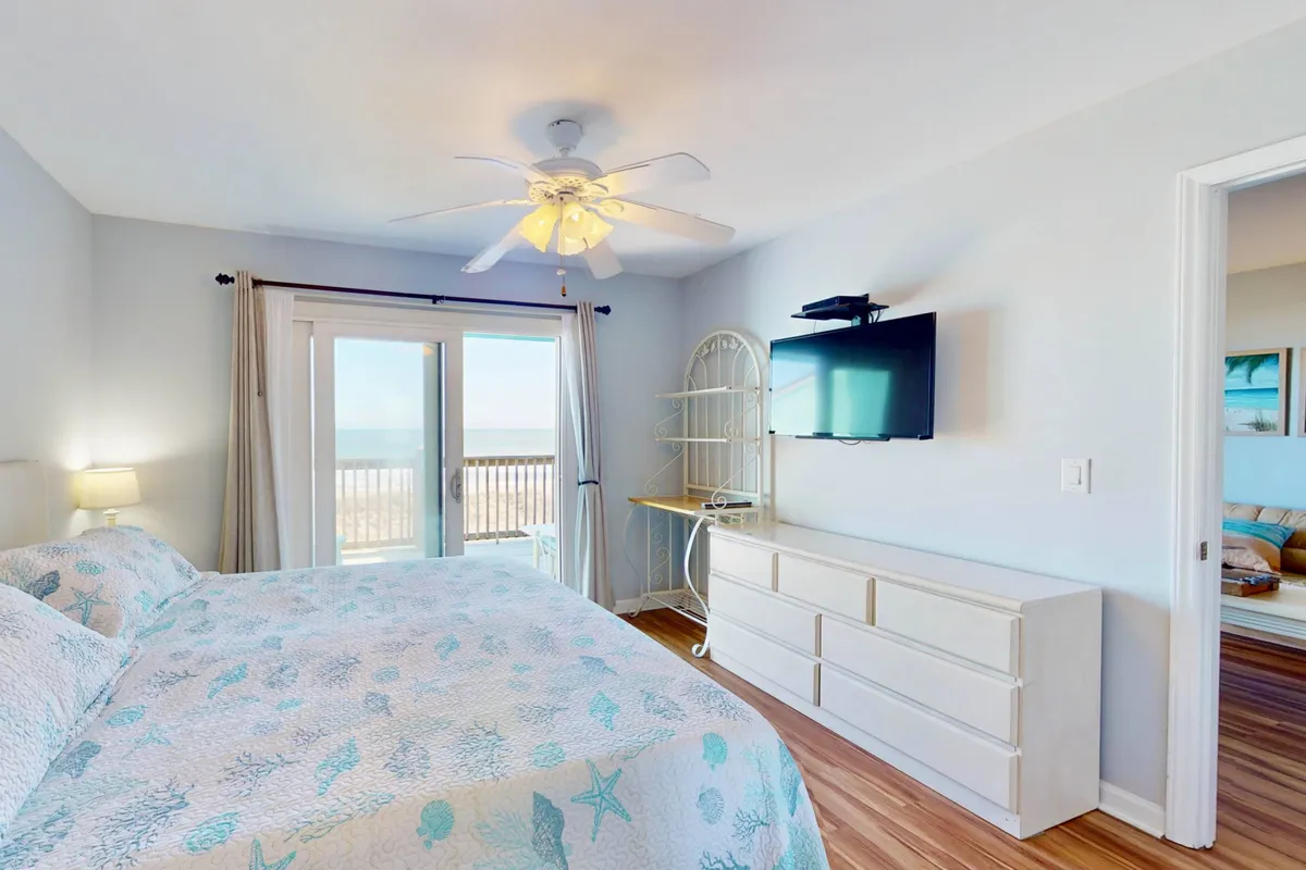 A calming bedroom featuring a sea-themed king bed, balcony doors opening to an ocean view, and a wall-mounted flat-screen TV. The gentle color palette and inviting textures evoke restfulness and privacy.