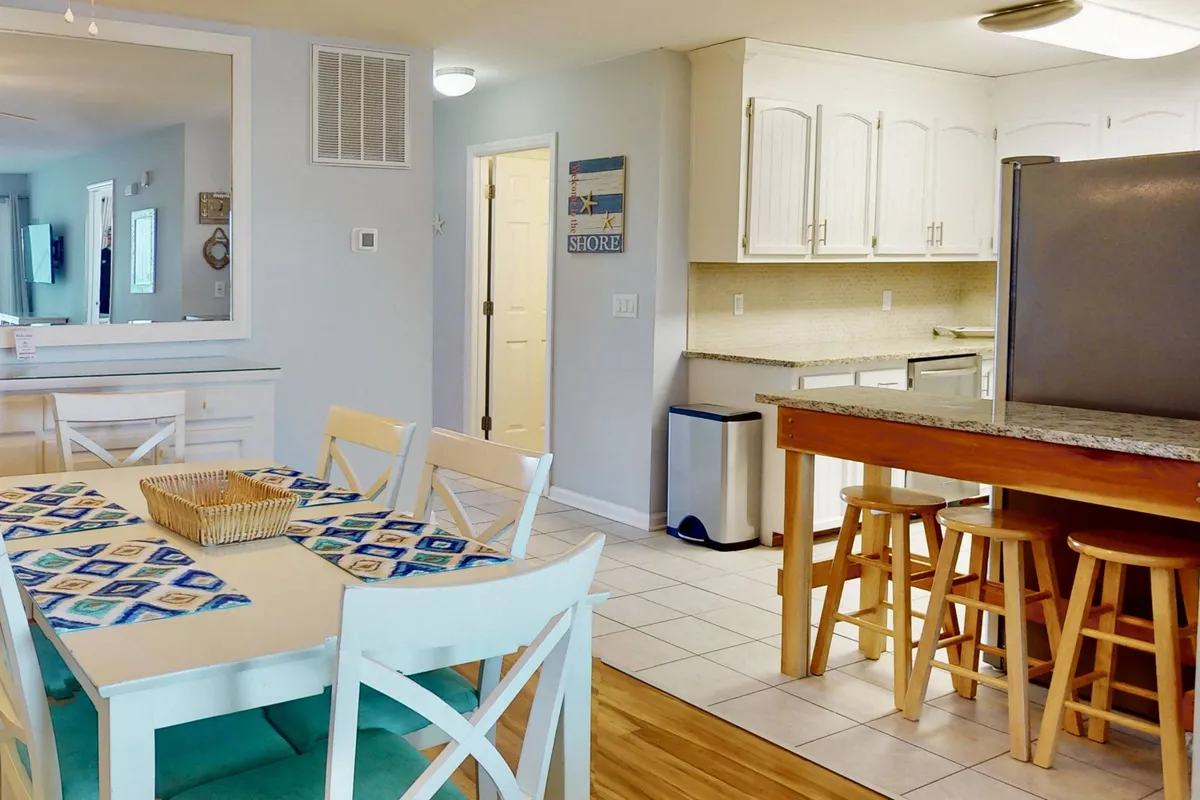 A bright and welcoming kitchen-dining space with a dining table adorned with a blue and beige table runner, a kitchen island with seating, and light-colored cabinetry. The setting is lively yet homey, ideal for cooking, baking, and socializing.