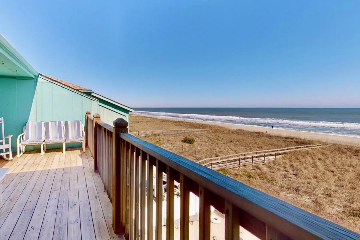 A serene exterior shot of a deck overlooking the beach, featuring a seating area and unobstructed ocean views. The bright scene invites sunbathing, reading, or gathering with friends for a barbecue or evening drinks.