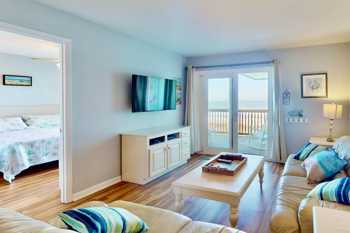 This image showcases a welcoming living room with comfortable leather sofas facing a large flat-screen TV. The space is bathed in natural light from sliding glass doors that lead directly to a balcony, granting a beautiful view of the ocean beyond.
