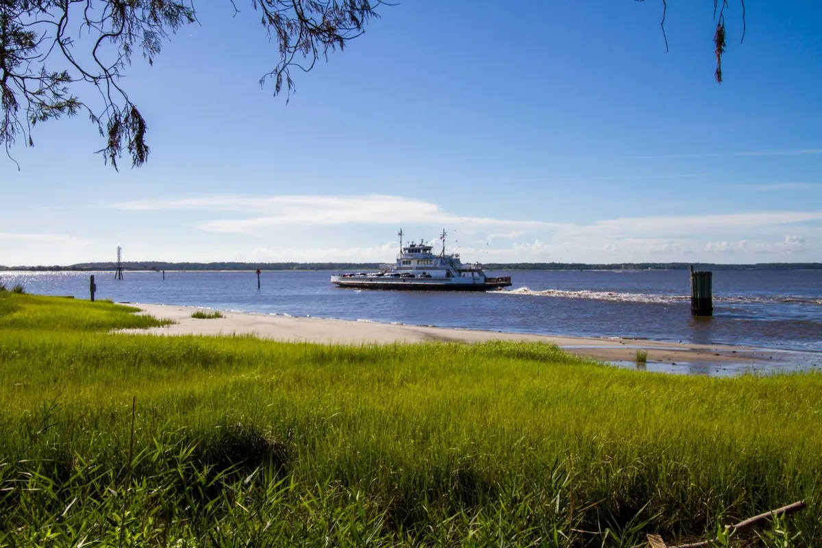 A peaceful riverside scene featuring a ferry boat gliding along the water, lush green grass on the banks, and wide-open skies. The setting is tranquil yet full of opportunity for boating, fishing, picnicking, and nature observation.