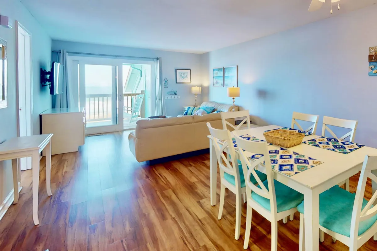 A bright, cheerful interior featuring a cozy living room sofa and a spacious dining table. The room is oriented toward sliding glass doors that offer sweeping views of the ocean, seamlessly blending indoor comfort with the natural beauty outside.