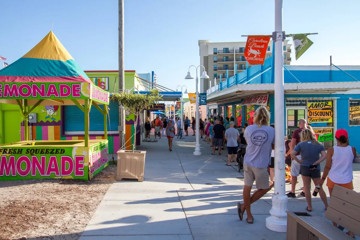 A bustling outdoor scene featuring a brightly colored lemonade stand surrounded by a crowd of people and vibrant storefronts. The area is full of energy with visitors exploring, chatting, and enjoying local refreshments.